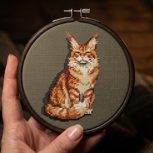以下が含まれることがあります： 木製の円形刺繍枠に収められた、オレンジ色のメインクーン猫のクロスステッチデザイン。猫はオレンジと白の毛並み、房毛のある尖った耳、長い白いひげが特徴で、無地のオリーブグリーンの布地を背景に配置されています。