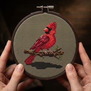 Puede incluir: Un bordado de punto de cruz de un cardenal rojo posado en una rama, dentro de un aro de madera redondo. El pájaro está detallado con varios tonos de rojo, y el fondo es verde apagado.
