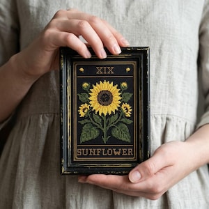 May include: A framed cross-stitch featuring a yellow sunflower with green leaves and buds against a black background. The frame has a distressed black finish with gold accents. The word "SUNFLOWER" is stitched at the bottom.