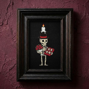 May include: Cross-stitch artwork of a skeleton in a top hat, holding roses and a box of skull chocolates. The framed piece has a black background and a dark wooden frame, ideal for Halloween decorations.