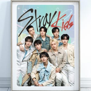 May include: A framed poster featuring the group Stray Kids. The image shows nine young men in various outfits, including denim jackets and neutral-toned jackets. The words "Stray Kids" are written in a stylized font across the top.