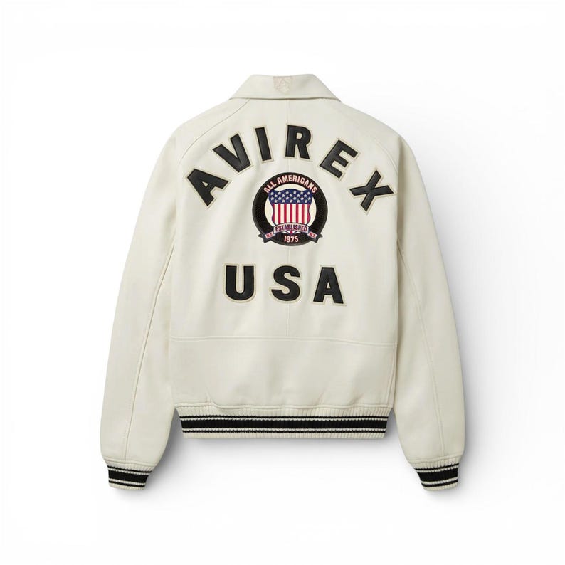 May include: Cream-colored leather jacket with the word "AVIREX" arched across the shoulders. Below, the word "USA" is displayed. A circular patch with the American flag and the words "ALL AMERICANS ESTABLISHED 1975" is centered above "USA". Black and white striped cuffs.
