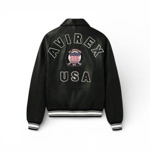 May include: Black leather jacket with white trim on the cuffs and hem. The back features the word "AVIREX" arched above a circular emblem with "ALL AMERICANS" and "1975" and the word "USA" below.