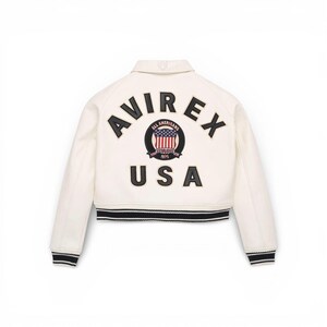 Puede incluir: Chaqueta bomber color crema con ribete a rayas blancas y negras. La parte trasera presenta la palabra "AVIREX" sobre un emblema circular con las palabras "ALL AMERICANS" y "USA". El emblema incluye un dise&ntilde;o de la bandera americana.