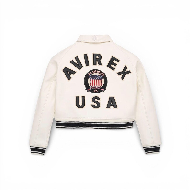 Puede incluir: Chaqueta bomber color crema con ribete a rayas blancas y negras. La parte trasera presenta la palabra "AVIREX" sobre un emblema circular con las palabras "ALL AMERICANS" y "USA". El emblema incluye un dise&ntilde;o de la bandera americana.