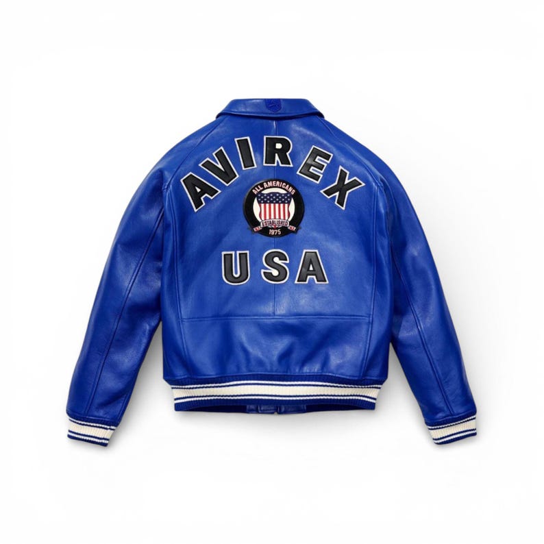 May include: A vibrant blue leather jacket with the word "AVIREX" arched across the back in white block letters. Below, the word "USA" is displayed. A circular emblem with "All Americans" and "1975" is centered above the "USA" text. The jacket has white and black striped cuffs.