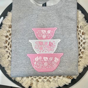 Puede incluir: Una sudadera gris con un diseño bordado de tres cuencos rosas y blancos anidados. Los cuencos presentan patrones florales y de bayas. La sudadera está hecha de un material suave y cómodo.