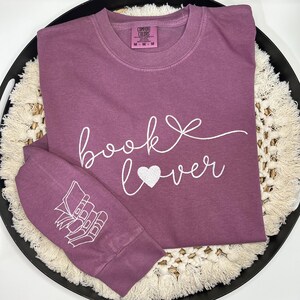 Op de afbeelding: Een mauvekleurig shirt met lange mouwen met de woorden "book lover" in witte letters, met een hart in plaats van het woord "love". De linkermouw heeft een witte afbeelding van gestapelde boeken. Het shirt heeft een ronde hals en een Comfort Colors-label.