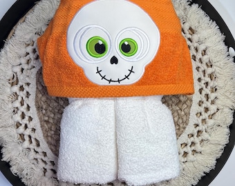 May include: An orange hooded towel with a white skull face design and green eyes. The towel is folded over a white towel, creating a Halloween-themed set. The set is displayed on a decorative tray.