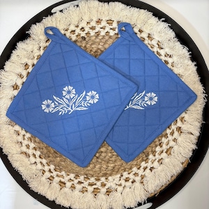 Puede incluir: Dos agarradores de cocina cuadrados y acolchados en color azul con un estampado floral blanco. Cada agarrador tiene un patrón de costura en forma de diamante y una pequeña trabilla de tela en la esquina superior para colgar. Tres flores blancas.