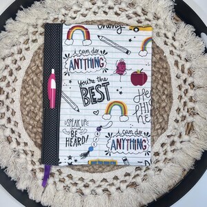 May include: A fabric notebook cover with a white background, featuring colorful illustrations of rainbows, pencils, and apples. Text includes "I can do ANYTHING" and "You're the BEST". A pink pen is in the side pocket. The cover is approximately 9 inches tall.