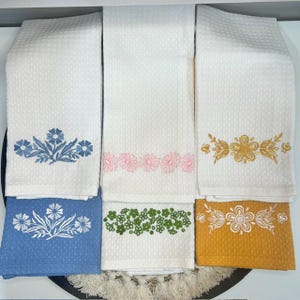 May include: A collection of six white waffle-weave kitchen towels with embroidered floral designs. The towels feature blue, pink, green, and gold floral patterns. Two blue and one yellow towel are visible at the bottom.