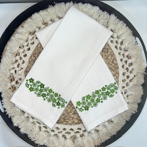 May include: Two white kitchen towels with a green floral embroidered border. The towels are folded and placed on a decorative woven tray with a black rim and fringe.