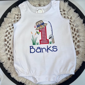 May include: A white sleeveless romper featuring an embroidered design of a red and white checkered number one, a tan fishing hat, a fishing rod, and green reeds. The name Banks is embroidered in blue below the graphic. The romper has button closures.