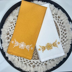 May include: Two decorative kitchen towels. One is mustard yellow with white floral embroidery, the other is white with gold floral embroidery. Both towels have a textured waffle weave and are folded neatly.