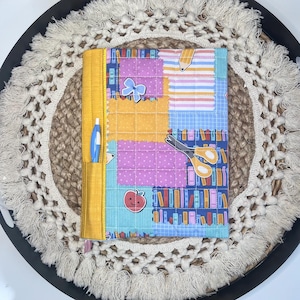 May include: A colorful, quilted notebook cover with a yellow spine and pen pocket. The cover features patchwork squares in various colors and patterns, including a blue bookshelf print, a red apple, and a blue bow. A blue pen is in the pocket.