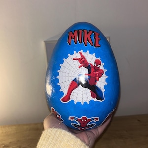 Puede incluir: Un objeto decorativo azul en forma de huevo con gráficos de Spider-Man. El superhéroe se representa en poses de acción con un fondo de telaraña. El nombre "MIKE" está escrito en letras rojas en la parte superior. También se ven logotipos adicionales de Spider-Man.