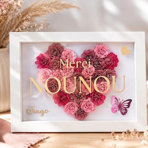Peut inclure: Un cadre vitrine rectangulaire blanc contenant un arrangement de fleurs en papier en forme de cœur dans des tons rose et bordeaux. La vitre porte l'inscription dorée Merci NOUNOU et Tiago, avec un petit cœur doré et un papillon rose.