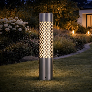 May include: A stainless steel cylindrical outdoor lamp. The lamp has a lattice design that allows light to shine through. It is positioned in a garden, with other lights in the background, creating a warm atmosphere. The lamp is approximately 100 cm tall.