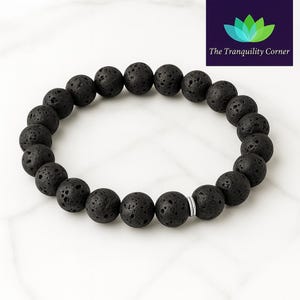 May include: Black lava stone bead bracelet. The bracelet features round, porous black beads strung together with a silver-colored spacer. The beads are evenly sized and have a textured surface. The background is a white marble-like surface.