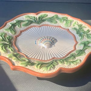 May include: A decorative ceramic plate with a scalloped edge. The plate features a central seashell design, surrounded by a radial pattern and green leaf accents. The rim is a terracotta color, and the plate has a cream-colored background.