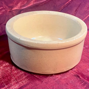 May include: A round, beige ceramic bowl with a slightly textured surface. The bowl has a wide rim and a simple, classic design. The interior is a lighter shade of beige. The bowl is suitable for serving or as a decorative piece.