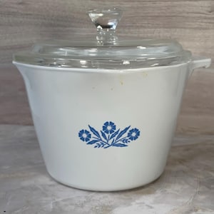 May include: White casserole dish with a clear glass lid and handle. The dish features a blue floral design on the front. The lid has a clear glass knob. The dish is likely vintage.