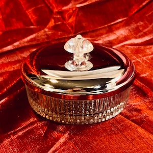 May include: A round, clear glass and silver-toned container with a lid and a decorative, mushroom-shaped handle. The container has a ribbed design on the sides and sits on a red velvet surface.