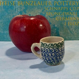 May include: A small Heise Bunzlauer Pottery spongeware schnapps cup with a 1 fl oz capacity, featuring a blue and green patterned design on a cream-colored base, positioned next to a large red apple with text overlaying the image.