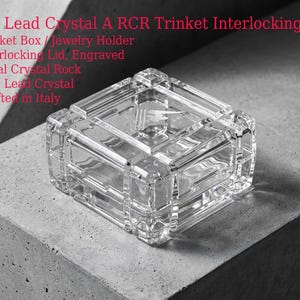 May include: A clear, square lead crystal trinket box with an interlocking lid. The box is made of 24% lead crystal and is designed to hold jewelry or small items. The text on the image says "24% Lead Crystal A RCR Trinket Interlocking Box".