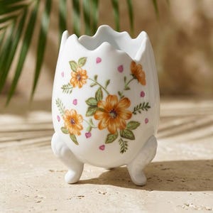 May include: A white ceramic egg-shaped vase with a scalloped rim, supported by three small feet. The vase is decorated with orange floral patterns and pink accents. The background features a blurred green palm leaf.