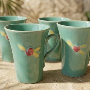 May include: Four turquoise ceramic mugs with handles. Each mug features a hand-painted design of a red rose with yellow leaves. The mugs have a flared shape and are set on a textured surface.