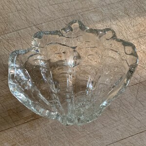 May include: A clear glass seashell-shaped bowl. The bowl has a textured surface and a scalloped edge. The bowl is empty and sits on a wooden surface. The bowl is likely used for decorative purposes.