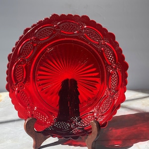 May include: A circular ruby red glass plate featuring a scalloped edge and a central sunburst pattern. The border is decorated with an embossed lattice and bead design. The plate is displayed upright on a dark brown wooden stand.