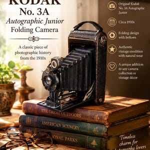 May include: A vintage Kodak No. 3A Autographic Junior folding camera with black bellows and a textured leather-like body. The camera features a metal lens assembly and a top handle, displayed on a stack of three antique books with visible text.