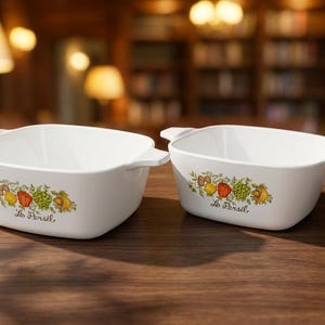 May include: Two white, square-shaped casserole dishes with handles. Each dish features a colorful floral and vegetable design with the text "Le Persil". The dishes are set on a wooden surface.