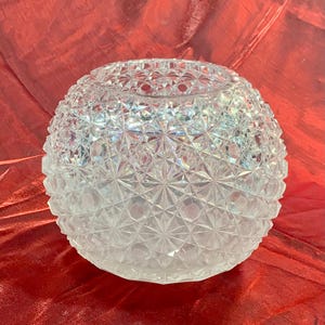 May include: A clear, spherical glass candle holder with a detailed, faceted surface. The intricate design creates a sparkling effect, reflecting light. The candle holder sits on a red surface.