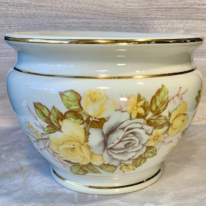 May include: A white ceramic planter with a gold rim and base. The planter is decorated with a floral pattern of yellow and white roses, green leaves, and small yellow flowers. The planter is a decorative item.