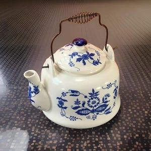 May include: A white ceramic teapot featuring a blue floral pattern, a matching lid with a blue knob, and a copper-colored metal wire handle with a coiled grip. The spout and body are decorated with intricate blue botanical designs.