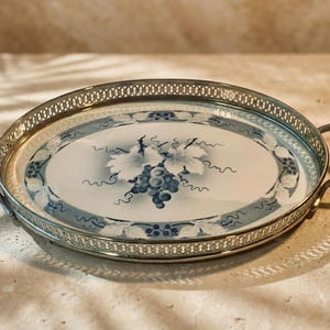 May include: Oval serving tray with a silver-colored metal rim and handles. The tray's interior features a white surface with a blue grape design. The rim has a decorative openwork pattern. The tray is suitable for serving food and drinks.