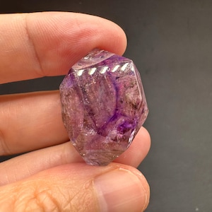 May include: A faceted, translucent purple crystal with internal dark inclusions and a polished surface. The gemstone displays varying shades of violet and clear areas, with sharp geometric edges and a multi-faceted cut that reflects light.