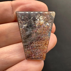 Peut inclure: Une pierre précieuse polie en forme de trapèze avec une surface chatoyante et irisée. La pierre présente un mélange de couleurs, dont l'or, le marron et des touches de rouge et de violet. La pierre est tenue dans une main.