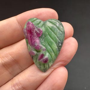 May include: A carved gemstone ornament featuring a deep magenta frog resting on a textured green heart-shaped leaf. The piece displays detailed relief work on the foliage and a smooth, polished surface on the frog, showcasing natural color variations.