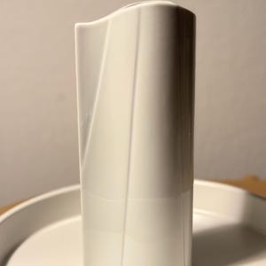 May include: A tall, white ceramic vase with a unique, curved design. The vase has a smooth, glossy finish and stands on a white, round tray. The vase's shape is rectangular with a slightly curved top and a subtle, angled detail on the front.