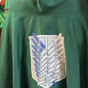 Puede incluir: Capa con capucha verde oscuro con un emblema blanco con alas azules y blancas. El texto "APR 6, 2026" está en la parte inferior. La capa está hecha de una tela suave.