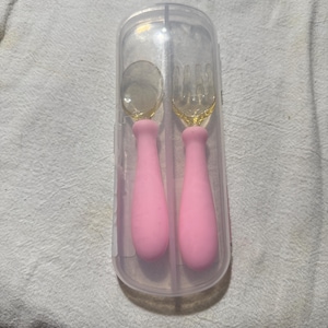 May include: A baby spoon and fork set in a clear plastic travel case. The spoon and fork have pink handles and clear, rounded eating surfaces. The case is rectangular with rounded edges. The set is designed for infant feeding.