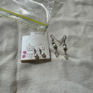 May include: Two pairs of earrings on a white surface. One pair is on a cardboard tag and features small, round, white pearls. The other pair has dangling pearls and clear crystal beads.