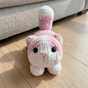 May include: A handmade crochet cat in pink and white. The cat has large black eyes, pink cheeks, and a pink and white striped tail. The cat is lying down on a wooden surface.