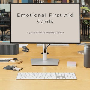 Könnte beinhalten: Ein Computermonitor zeigt den Text "Emotional First Aid Cards" und "A 90-card system for returning to yourself." Der Monitor steht auf einem Holzschreibtisch mit Tastatur, Tacker und anderem Büromaterial.