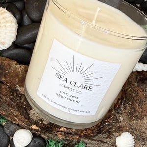 May include: A large glass candle with a creamy wax fill, labeled "SEA CLARE CANDLE CO." and "NEWPORT, RI." The candle is hand-poured with coconut soy wax and rests on a piece of driftwood, surrounded by black stones, seashells, and green moss.
